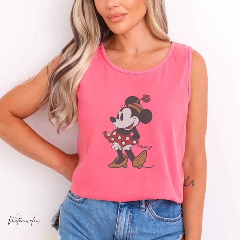 Peut inclure: Un d&eacute;bardeur rose avec un motif de Minnie Mouse. Le personnage de dessin anim&eacute; porte une robe &agrave; pois rouges, des chaussures marron et un chapeau avec une fleur. Le mot "Disney" est imprim&eacute; sous le motif.