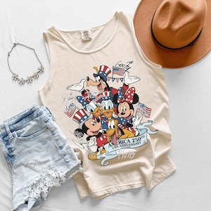 May include: An ivory tank top featuring a graphic print of Disney characters including Mickey Mouse, Minnie Mouse, Donald Duck, Daisy Duck, Goofy, and Pluto wearing patriotic hats. The text reads AMERICA 250TH USA ANNIVERSARY 1776-2026.