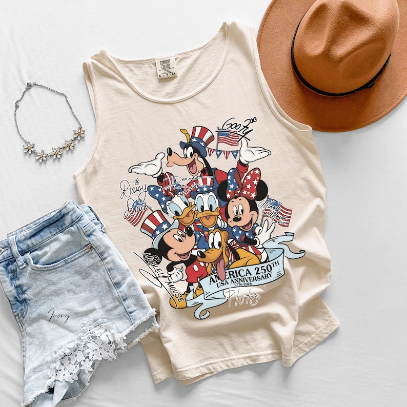 May include: An ivory tank top featuring a graphic print of Disney characters including Mickey Mouse, Minnie Mouse, Donald Duck, Daisy Duck, Goofy, and Pluto wearing patriotic hats. The text reads AMERICA 250TH USA ANNIVERSARY 1776-2026.