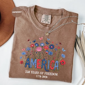 May include: A brown Comfort Colors t-shirt with a floral design and the word "AMERICA" in blue and red. The shirt also features the text "250 YEARS OF FREEDOM 1776-2026". A silver necklace with a pendant is draped across the shirt.