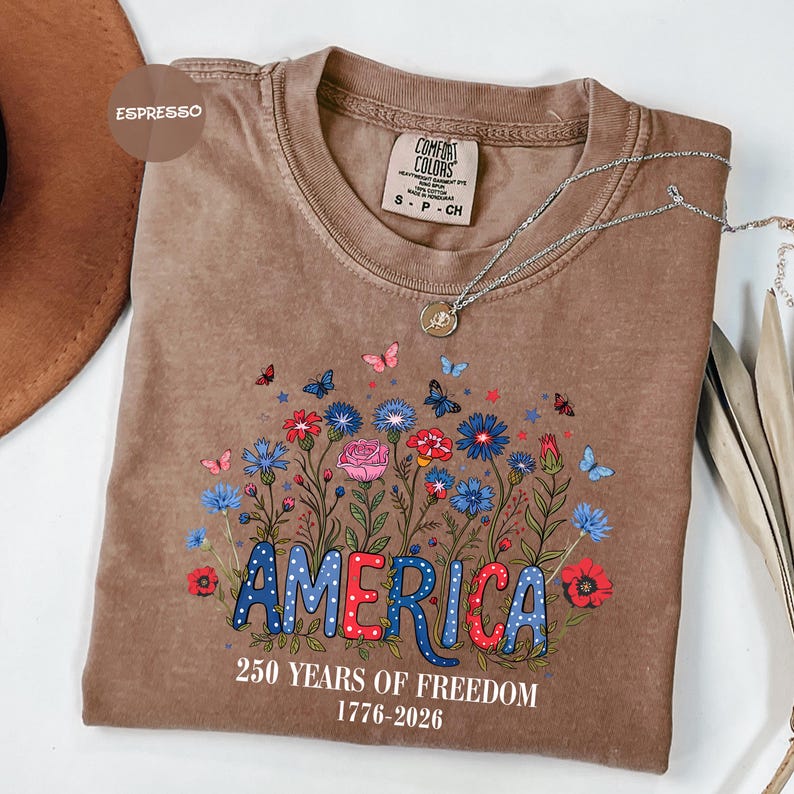 May include: A brown Comfort Colors t-shirt with a floral design and the word "AMERICA" in blue and red. The shirt also features the text "250 YEARS OF FREEDOM 1776-2026". A silver necklace with a pendant is draped across the shirt.