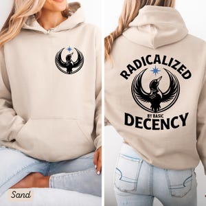 May include: Sand-colored hoodie with a front pocket and a hood. The front features a black graphic of a bird within a circle and a star. The back of the hoodie displays the text "RADICALIZED BY BASIC DECENCY" around a similar bird graphic.