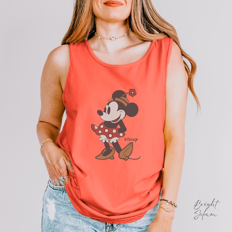 Peut inclure: D&eacute;bardeur corail avec un motif de Minnie Mouse. Le personnage de dessin anim&eacute; est repr&eacute;sent&eacute; dans une pose classique, portant une robe rouge &agrave; pois blancs et des chaussures marron. Le logo Disney est visible.
