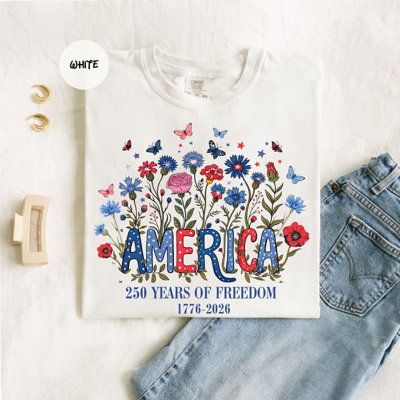 May include: White t-shirt with a floral design and the word "AMERICA" in red, white, and blue. The shirt features a floral arrangement with butterflies and the text "250 YEARS OF FREEDOM 1776-2026".