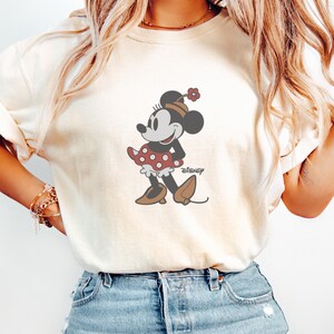 Peut inclure: T-shirt cr&egrave;me avec un motif de Minnie Mouse. Minnie porte une robe &agrave; pois rouges, des chaussures marron et un chapeau fleuri. Le logo Disney est imprim&eacute; en dessous. Le mot "Ivory" est &eacute;crit en cursive.