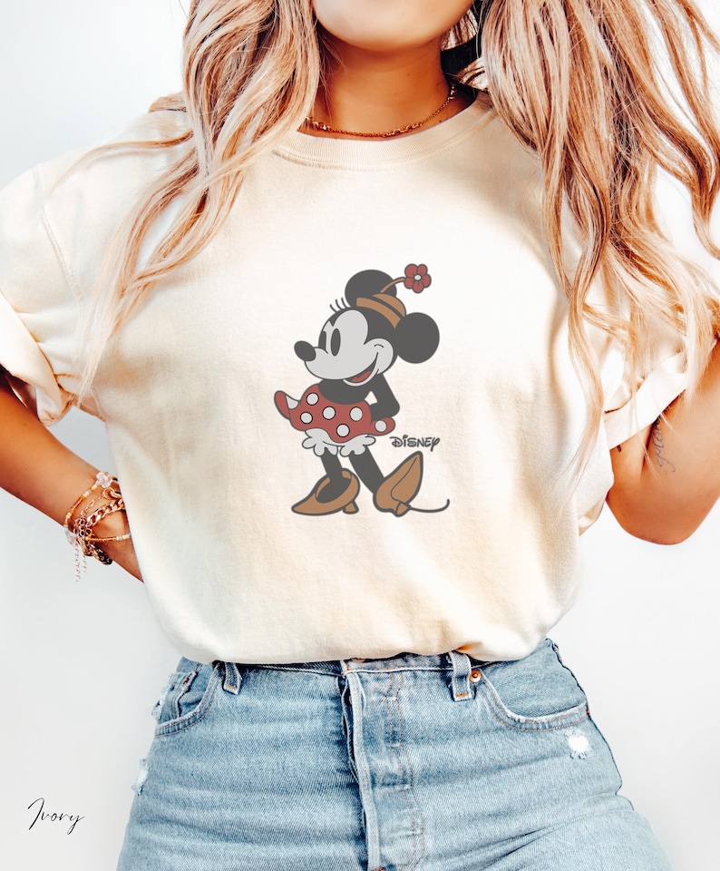 Peut inclure: T-shirt cr&egrave;me avec un motif de Minnie Mouse. Minnie porte une robe &agrave; pois rouges, des chaussures marron et un chapeau fleuri. Le logo Disney est imprim&eacute; en dessous. Le mot "Ivory" est &eacute;crit en cursive.