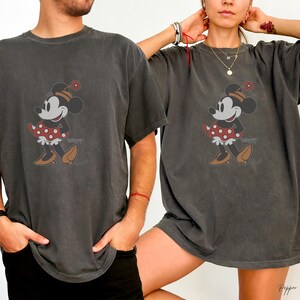Peut inclure: T-shirts gris fonc&eacute; avec un motif de Minnie Mouse. Le personnage de dessin anim&eacute; porte une robe &agrave; pois rouges, des chaussures marron et un chapeau fleuri. Le mot "Disney" est imprim&eacute; en dessous.