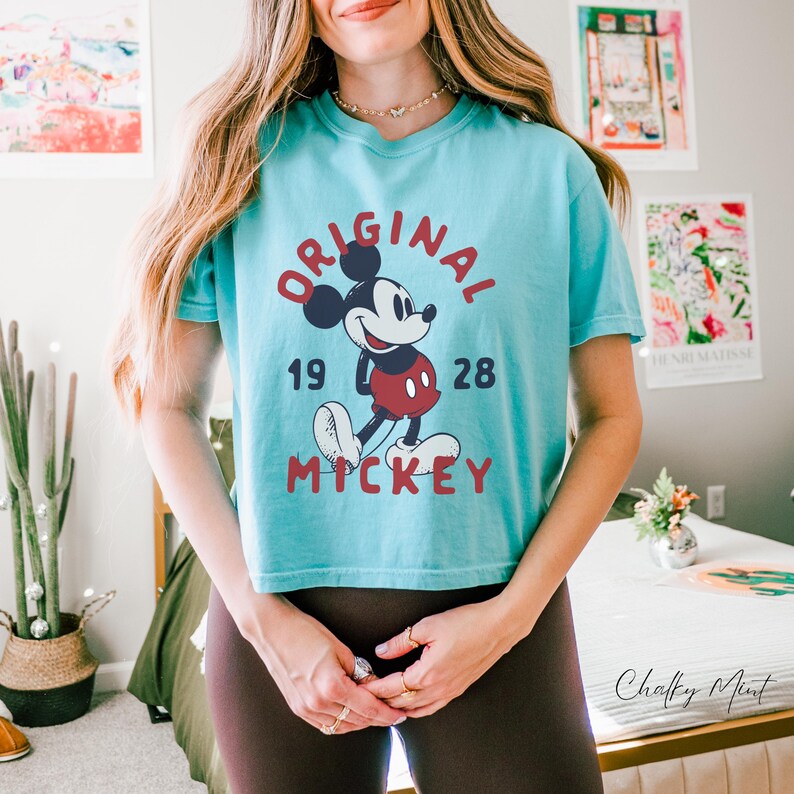 Puede incluir: Una camiseta de manga corta de color turquesa claro con un gr&aacute;fico de Mickey Mouse en el centro. El texto "ORIGINAL" est&aacute; arqueado encima, "19" y "28" est&aacute;n a los lados, y "MICKEY" est&aacute; impreso debajo en una fuente roja oscura y negrita.