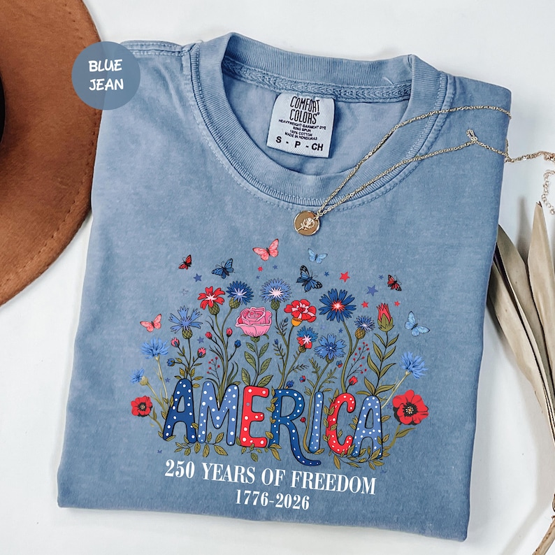 May include: A blue jean colored t-shirt with a floral design and the word "AMERICA" in a patriotic font. The shirt also features the text "250 YEARS OF FREEDOM 1776-2026". A gold necklace is draped across the shirt.