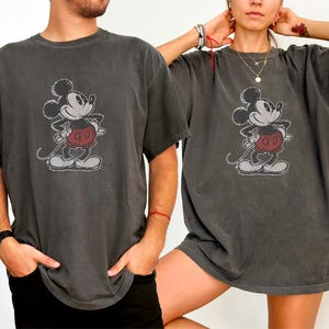 May include: A charcoal gray short-sleeve t-shirt featuring a distressed graphic print of Mickey Mouse on the front. The character is depicted in black and white with red shorts, standing with hands on hips in a classic pose on the center chest.