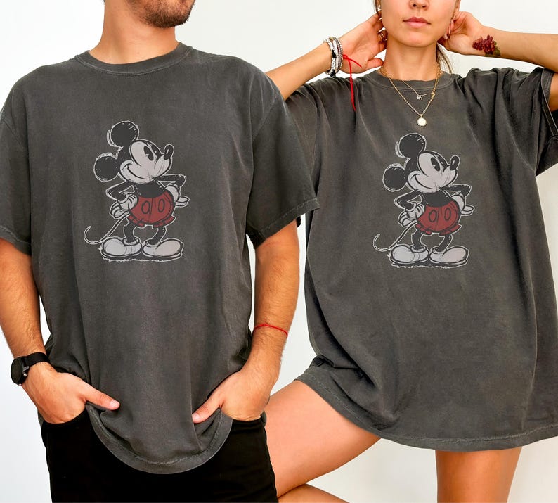 May include: A charcoal gray short-sleeve t-shirt featuring a distressed graphic print of Mickey Mouse on the front. The character is depicted in black and white with red shorts, standing with hands on hips in a classic pose on the center chest.
