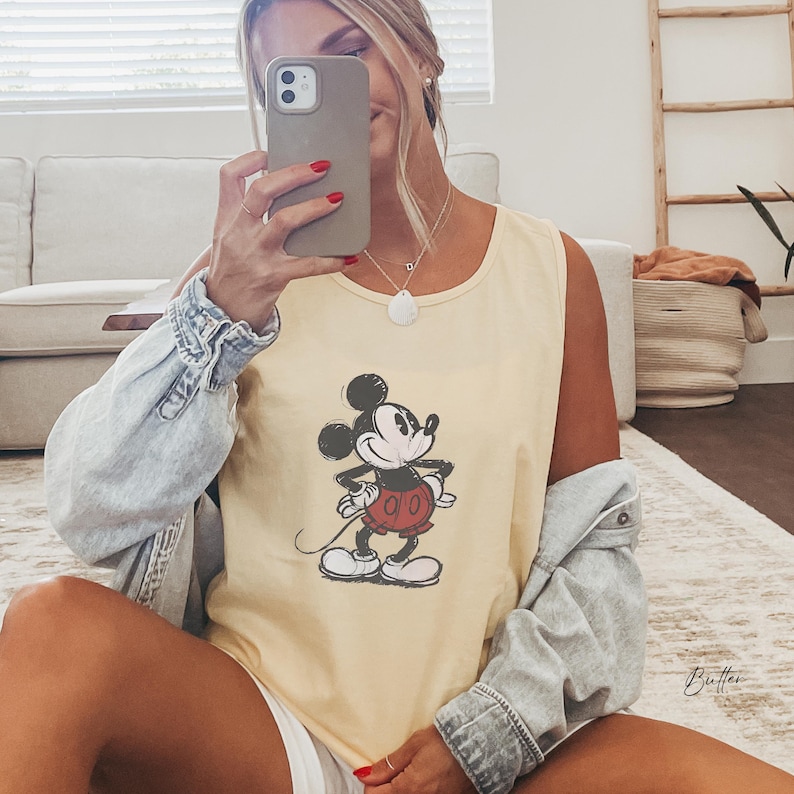 May include: A pale yellow sleeveless tank top featuring a vintage-style graphic of Mickey Mouse with black ears, a black body, and red shorts. The tank top is worn with a light blue denim jacket draped over the shoulders, creating a casual layered look.