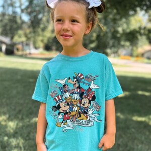May include: A seafoam green short-sleeve t-shirt featuring a graphic print of Disney characters including Mickey Mouse, Minnie Mouse, Donald Duck, Daisy Duck, Goofy, and Pluto wearing patriotic hats, with the text America 250th USA Anniversary.
