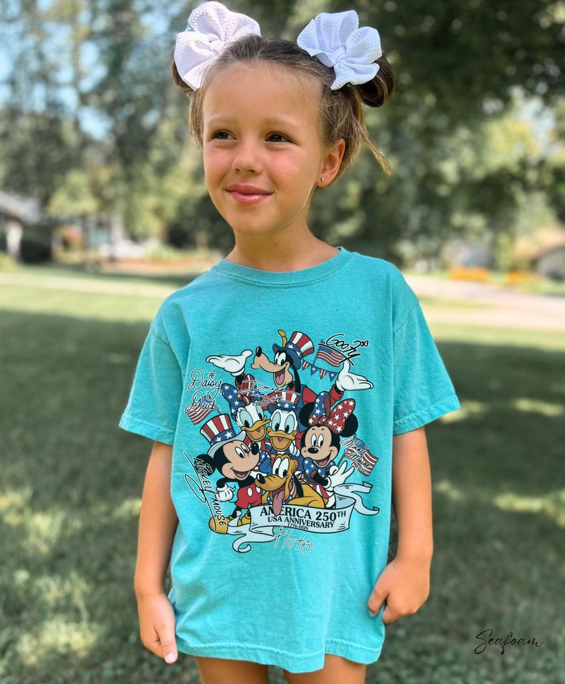 May include: A seafoam green short-sleeve t-shirt featuring a graphic print of Disney characters including Mickey Mouse, Minnie Mouse, Donald Duck, Daisy Duck, Goofy, and Pluto wearing patriotic hats, with the text America 250th USA Anniversary.