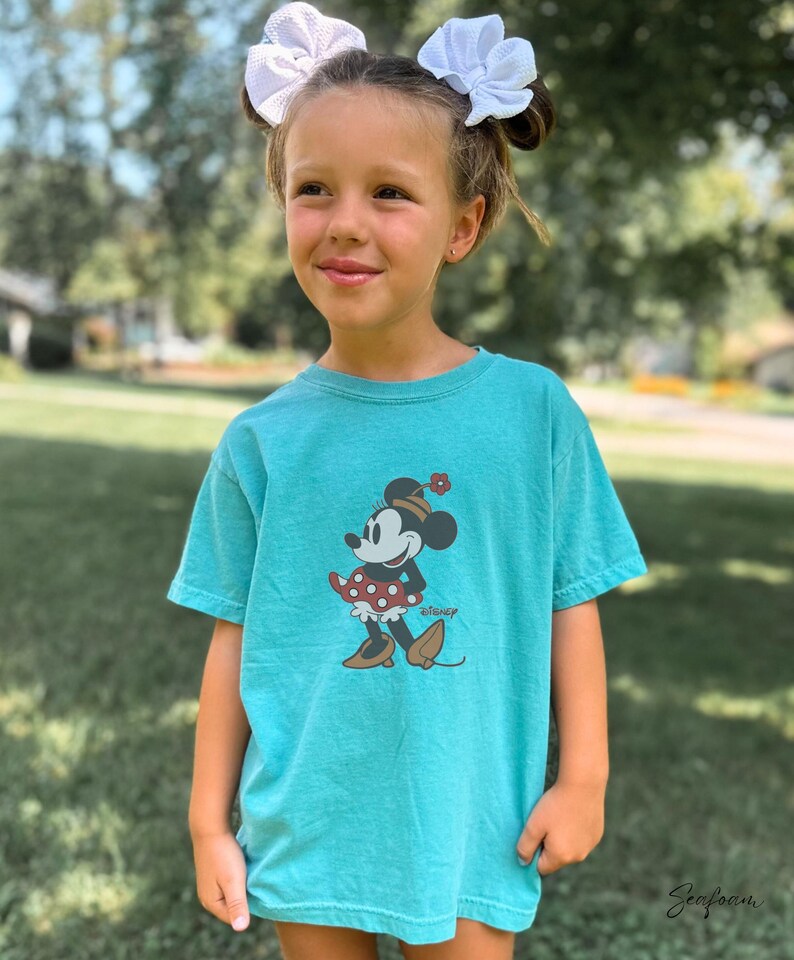 Peut inclure: Un t-shirt turquoise avec un motif de Minnie Mouse. Le personnage de dessin anim&eacute; porte une jupe &agrave; pois rouges, un chapeau avec une fleur et des chaussures marron. Le t-shirt a un col rond et des manches courtes. Le mot "Disney" est imprim&eacute; en dessous.