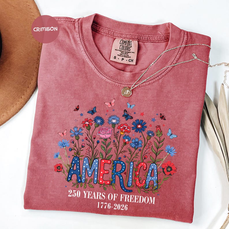 May include: Crimson-colored t-shirt with the word "AMERICA" in a patriotic design, surrounded by flowers and butterflies. The shirt also features the text "250 YEARS OF FREEDOM 1776-2026". A silver necklace with a pendant is draped across the shirt.