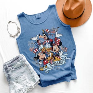 May include: A blue sleeveless tank top featuring a graphic print of Disney characters including Mickey Mouse, Minnie Mouse, Donald Duck, Daisy Duck, Goofy, and Pluto wearing patriotic hats. The text reads America 250th USA Anniversary 1776-2026.