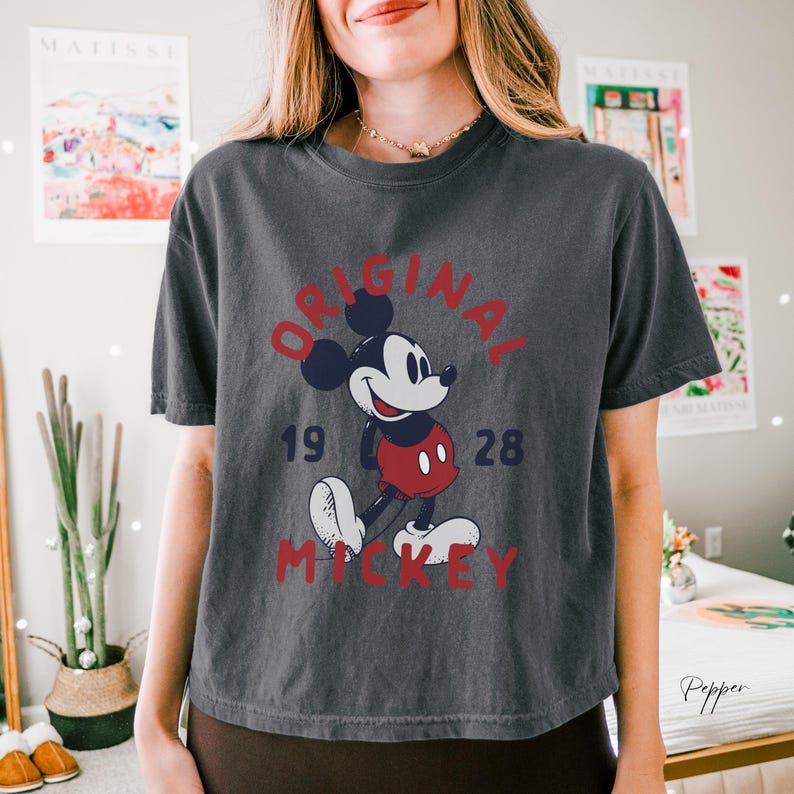 Puede incluir: Una camiseta de manga corta gris carb&oacute;n con un gr&aacute;fico de Mickey Mouse con pantalones cortos rojos y zapatos blancos. El texto "ORIGINAL" est&aacute; arqueado en rojo arriba, con "19" y "28" a los lados y "MICKEY" impreso en rojo debajo.