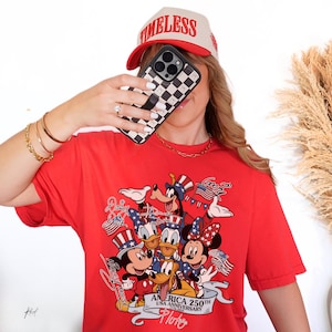 May include: A bright red short-sleeved t-shirt featuring a graphic print of various cartoon characters wearing patriotic hats. The design includes the text "AMERICA 250TH USA ANNIVERSARY 1776-2026" along with individual character names written in script.