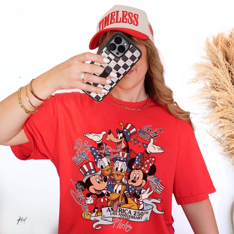 May include: A bright red short-sleeved t-shirt featuring a graphic print of various cartoon characters wearing patriotic hats. The design includes the text "AMERICA 250TH USA ANNIVERSARY 1776-2026" along with individual character names written in script.