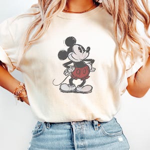 May include: An ivory short-sleeved t-shirt featuring a centered graphic illustration of Mickey Mouse in a classic pose. The character is depicted in black, white, and red tones with a distressed, vintage-style print texture on the fabric.