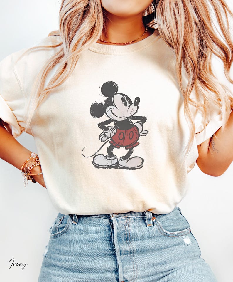 May include: An ivory short-sleeved t-shirt featuring a centered graphic illustration of Mickey Mouse in a classic pose. The character is depicted in black, white, and red tones with a distressed, vintage-style print texture on the fabric.