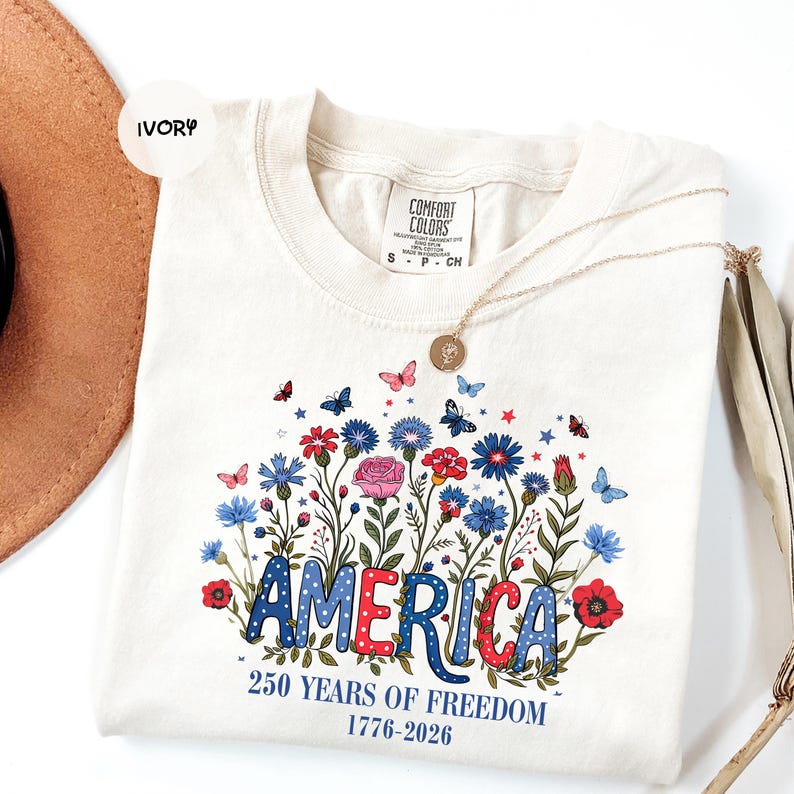 May include: Ivory-colored t-shirt with a floral design and the word "AMERICA" in red, white, and blue. The shirt also features the text "250 YEARS OF FREEDOM 1776-2026". A gold necklace and a brown hat are also visible.