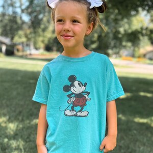 May include: A seafoam green short-sleeve t-shirt featuring a black and white graphic print of Mickey Mouse wearing red shorts. The shirt has a crew neckline and a relaxed fit. The word Seafoam is printed in a cursive script in the bottom corner.
