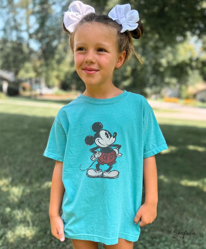 May include: A seafoam green short-sleeve t-shirt featuring a black and white graphic print of Mickey Mouse wearing red shorts. The shirt has a crew neckline and a relaxed fit. The word Seafoam is printed in a cursive script in the bottom corner.