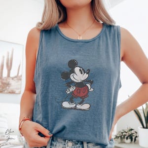 May include: A blue sleeveless tank top featuring a distressed graphic print of Mickey Mouse in the center. The character is depicted in a classic pose with black ears, a black body, red shorts, and white gloves and shoes, set against the blue fabric.
