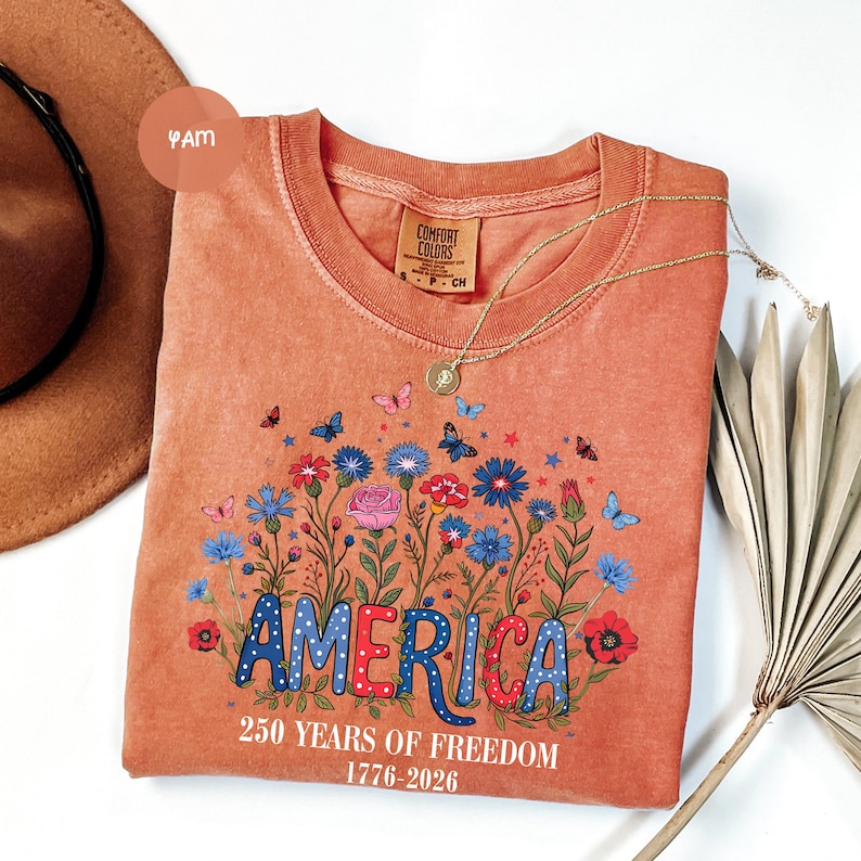 May include: Orange Comfort Colors t-shirt with a floral design and the word "AMERICA" in red, white, and blue. The shirt also features the text "250 YEARS OF FREEDOM 1776-2026". A gold necklace and a brown hat are also visible.