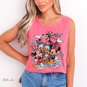May include: A bright pink sleeveless tank top featuring a graphic print of various cartoon characters wearing patriotic red, white, and blue hats. The text reads "AMERICA 250TH USA ANNIVERSARY 1776-2026" along with names of the characters in script font.