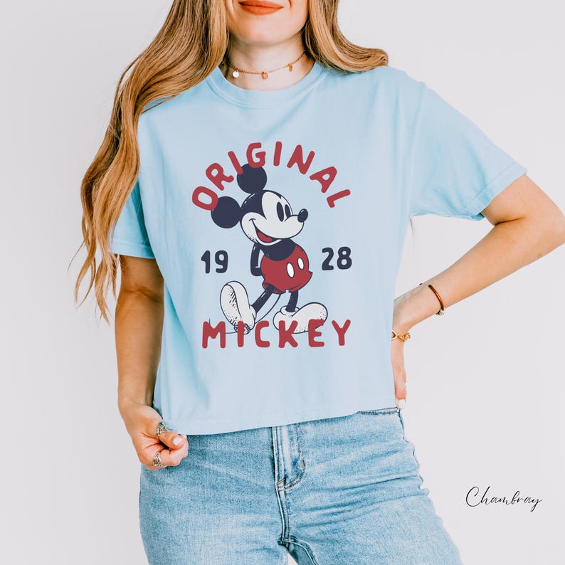 Puede incluir: Una camiseta de manga corta de color azul claro con un gr&aacute;fico de Mickey Mouse en el centro. El texto "ORIGINAL" est&aacute; arqueado sobre el personaje, "19" y "28" est&aacute;n a los lados, y "MICKEY" est&aacute; impreso debajo en una fuente roja.