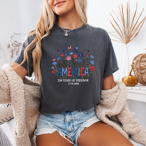 May include: Dark gray t-shirt with the word "AMERICA" in a floral design. The shirt also features the text "250 YEARS OF FREEDOM 1776-2026". The design includes red, white, and blue flowers and butterflies.