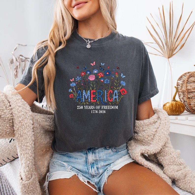 May include: Dark gray t-shirt with the word "AMERICA" in a floral design. The shirt also features the text "250 YEARS OF FREEDOM 1776-2026". The design includes red, white, and blue flowers and butterflies.