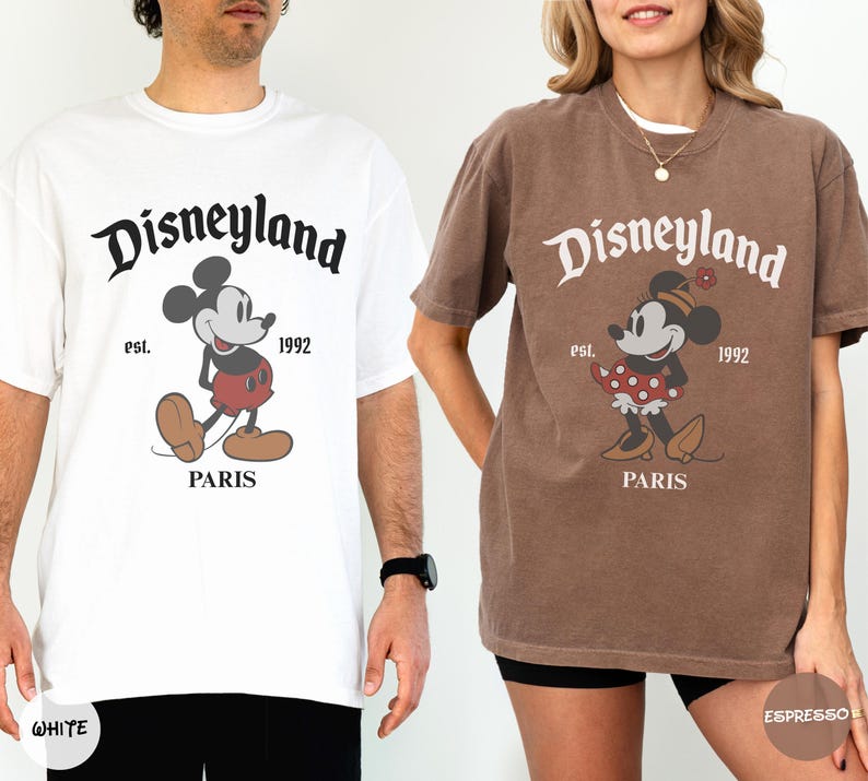 May include: Two t-shirts, one white and one brown, featuring a Disneyland Paris graphic. The graphic includes the Disneyland logo, Mickey Mouse, and the text "est. 1992 PARIS". The white shirt has a black graphic, and the brown shirt has a red and white graphic.