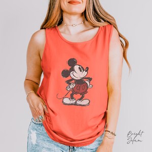 May include: A bright salmon-colored sleeveless tank top featuring a centered, distressed-style graphic print of Mickey Mouse in black and white. The character is depicted standing with hands on hips, wearing shorts, and shoes in a classic cartoon style.