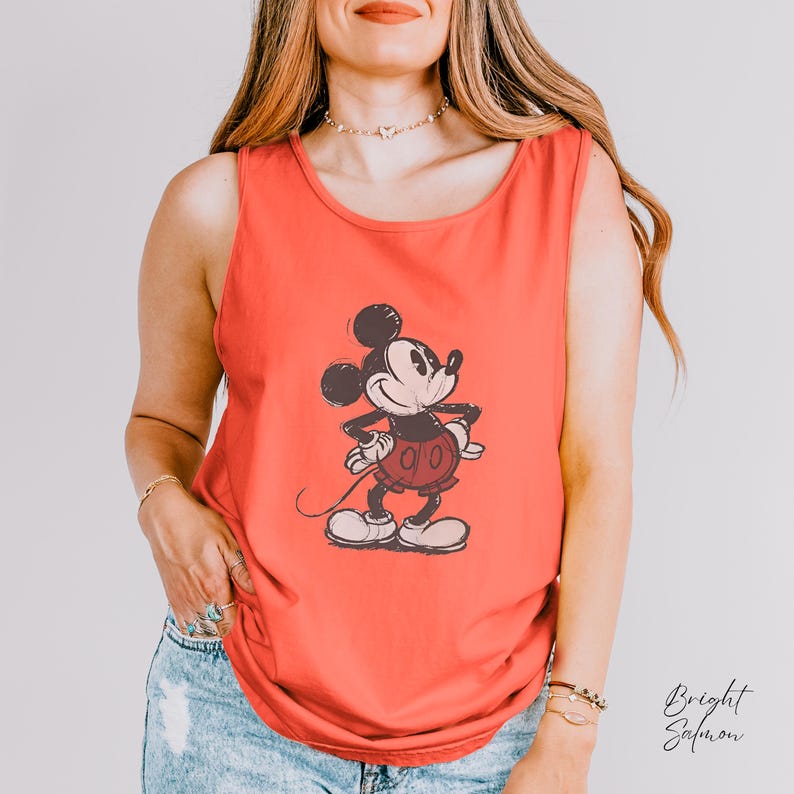 May include: A bright salmon-colored sleeveless tank top featuring a centered, distressed-style graphic print of Mickey Mouse in black and white. The character is depicted standing with hands on hips, wearing shorts, and shoes in a classic cartoon style.