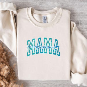 May include: Cream-colored sweatshirt with the word "MAMA" in a teal, block letter design. The word "mama" is written in a script font underneath, with a small blue heart. The sweatshirt has a crew neck and long sleeves.