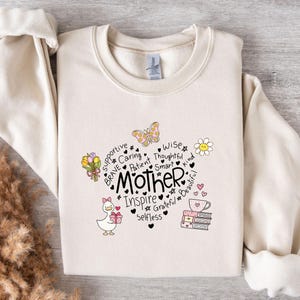 May include: Cream-colored sweatshirt with the word "Mother" in the center, surrounded by words like "supportive" and "brave." Adorable illustrations of a duck, flowers, and a teacup add charm. A perfect gift for Mother's Day.