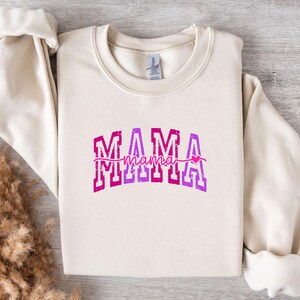 May include: A cream-colored sweatshirt with the word "MAMA" in a pink and purple ombre design. The word "mama" is written in a cursive font underneath, with a small pink heart. The sweatshirt has long sleeves.