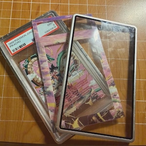 May include: A 1999 Pokemon Jungle Pikachu trading card in a PSA graded plastic slab, layered with a decorative card frame featuring a pink and purple sky illustration, and a clear rectangular display frame with a black border.