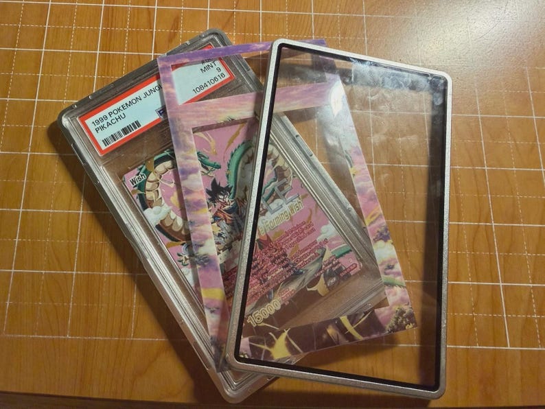 May include: A 1999 Pokemon Jungle Pikachu trading card in a PSA graded plastic slab, layered with a decorative card frame featuring a pink and purple sky illustration, and a clear rectangular display frame with a black border.