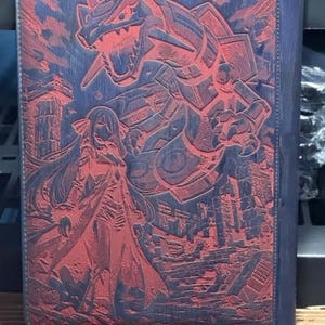 May include: A blue and red card binder featuring a detailed embossed design. The cover showcases a large dragon-like creature and a female figure, set against a cityscape. The binder has a zipper closure and a blue border.