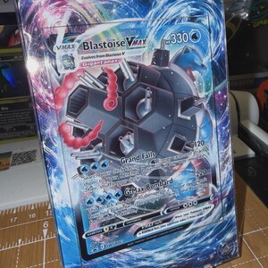 May include: A Blastoise VMAX Pokémon card, featuring a blue and black illustration of the character. The card has a holographic background with the text "Grand Falls" and "G-Max Bombard". The card is displayed in a clear acrylic holder.