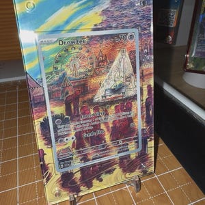 May include: A framed Drowzee Pokémon card. The card features a colorful illustration of a carnival scene with a Ferris wheel and a tent. The card has the text "Forced Sleep" and "Gentle Slap". The frame is clear and held up by a clear stand.