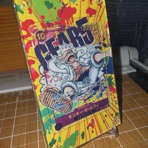 May include: A colorful card featuring the character Monkey D. Luffy from the anime series One Piece. The card has a yellow background with green and red paint splatters. The text "GEARS" and "12000" are visible.