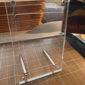 May include: A clear acrylic paper cutter with a silver metal guide and blade. The cutter is on a wooden surface with a grid pattern. A stack of brown envelopes is visible behind the cutter.