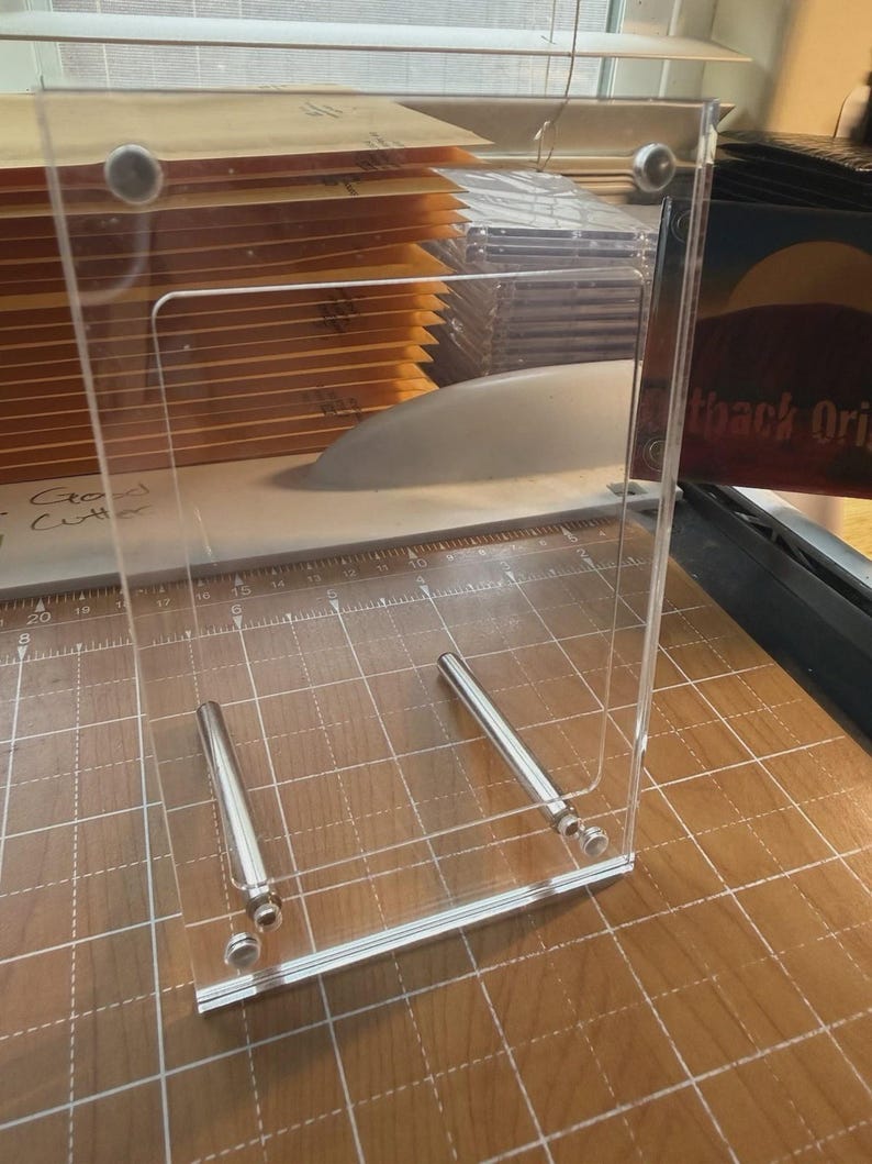 May include: A clear acrylic paper cutter with a silver metal guide and blade. The cutter is on a wooden surface with a grid pattern. A stack of brown envelopes is visible behind the cutter.