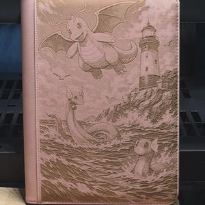 May include: Pink card binder featuring a detailed engraved design of a Dragonite flying above the ocean, a lighthouse, and other Pokemon. The binder has a light pink cover and spine.